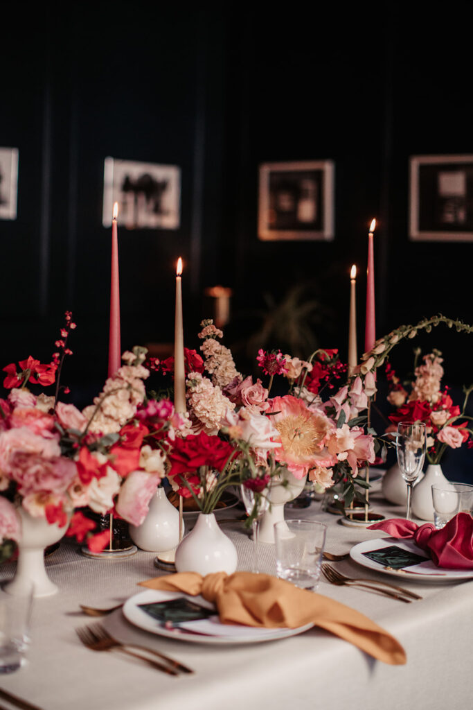 A luxury wedding table with lots of white vases with red, pink and blush flowers, tapered candles and napkins alternating in colour